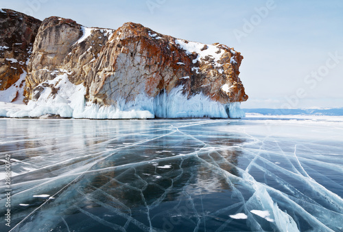 Beautiful winter landscape of the frozen Baikal Lake. Endless fields of blue slippery ice with cracks near the red rocks of Olkhon Island. View on the Strait of Small Sea on a sunny cold day