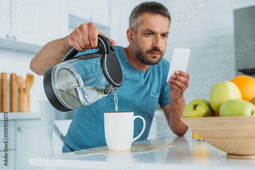 selective focus of man overfilling cup with water while sitting at kitchen table and using smartphone