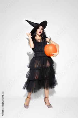 Young brunette woman in black hat and costume on white background. Attractive caucasian female model. Halloween, black friday, cyber monday, sales, autumn concept. Copyspace. Holds pumpking.