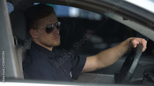 Confident policeman sitting in patrol car and wearing sunglasses, ready for work