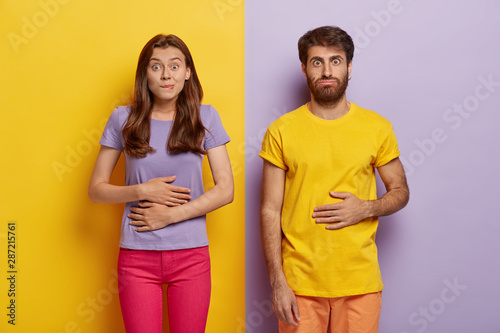 Young female and male touch stomaches, feel satiety after eating hearty meal, bites lips, have good appetite, dressed in casual outfit, pose against yellow and purple wall. Pleasant feeling in belly