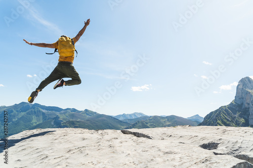 Young Man Jumping on Top of a Mountain Wearing Yellow Backpack.