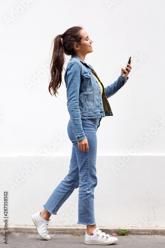 Full length young woman with brown hair walking and looking at mobile phone by white wall
