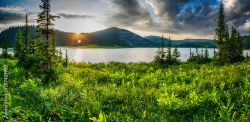 Beautiful mountain lake with pine trees and green grass on shore in front of hills during sunset, Ergaki national park, Siberia, Russia
