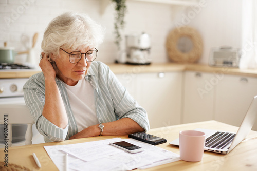 People, age, technology and finances. Depressed unhappy retired woman paying domestic bills online, trying hard to make both ends meet, sitting at kitchen table, surrounded with papers, using gadgets