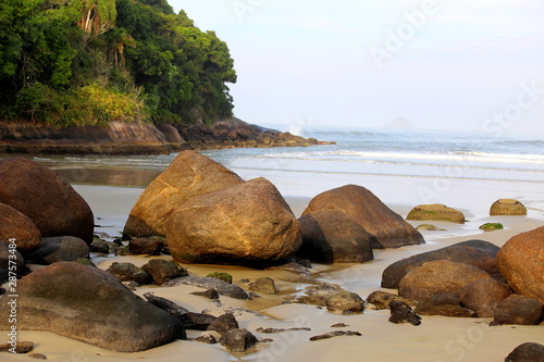 Beach with rocks on the sand. Calm waters and ocean view until the horizon. Hill with green vegetation on the left. Sao Lourenco beach, Bertioga, Brazil,