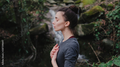 Young woman practicing breathing yoga pranayama outdoors in moss forest on background of waterfall. Unity with nature concept.