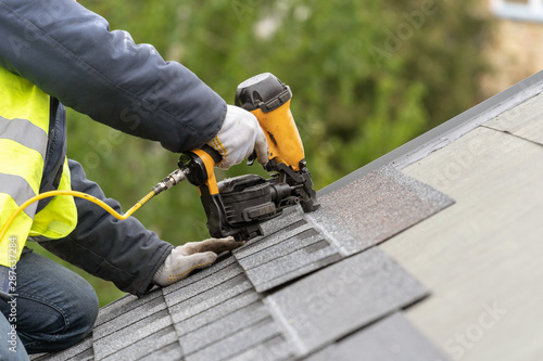 Workman using pneumatic nail gun install tile on roof of new house under construction