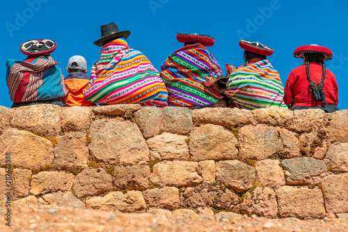 A group of Quechua indigenous women in traditional clothing and a young boy sitting and chatting on an ancient Inca wall in the archaeological site of Chinchero in the region of Cusco city, Peru.
