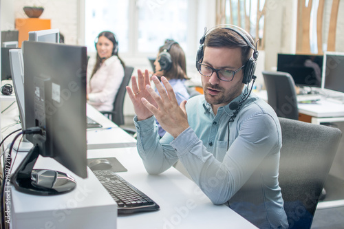 Young handsome male technical support agent trying to explain something to a client while using hands-free headset at call center