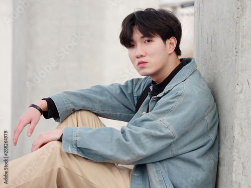 Portrait of a handsome Chinese young man in jeans sitting on ground and looking at camera.