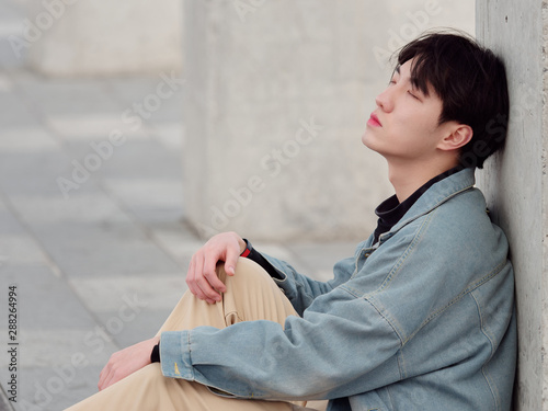 Portrait of a handsome Chinese young man sitting on ground and leaning against wall with eyes closed, tired young man have a little break.