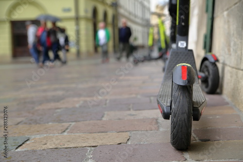 dwie hulajnogi elektryczne zaparkowane blisko budynku w centrum miesta, w tle idący ludzie