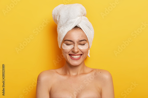 Headshot of satisfied beautiful naked woman applies white patches under eyes to reduce dryness, has pampering session, plumps up skin, wears white towel on head after shower, isolated on yellow wall