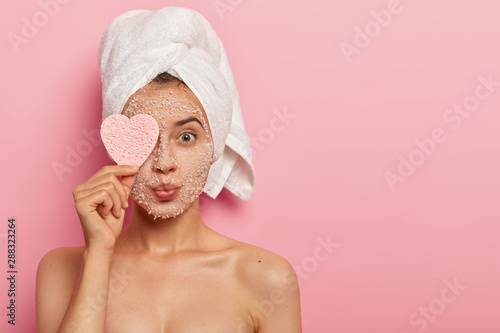 Reducing pores and cleansing concept. Attractive female applies sea salt mask on face, has luxurious feelings from beauty treatments, covers eye with heart shaped sponge, pampers complexion.