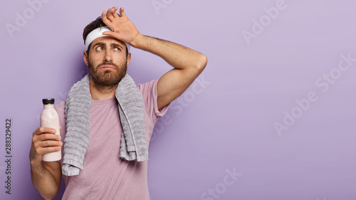 Exhausted athlete man feel thirsty and tired after hard cardio workout, wipes sweat on forehead, holds bottle of cold water, wears t shirt, towel around neck, relaxes after training, isolated