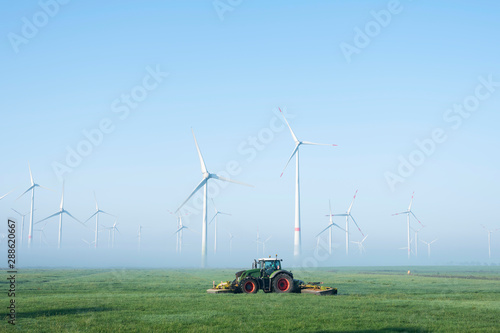 farmer mows grass near wind turbine farm in oastfriesland on misty summer morning