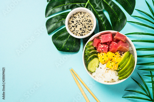 Tuna poke bowl with rice, avocado, mango and cucumber on blue.