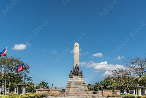 Manila, Philippines - March 5, 2019: Wide landscape shot of Obelisk with bronze statues of Jose Rizal stands on brown stone pedestal under blue sky in Rizal Park. Flags and green foliage.