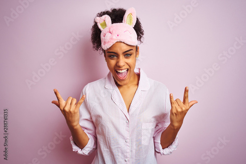 Young african american woman wearing pajama and mask over isolated pink background shouting with crazy expression doing rock symbol with hands up. Music star. Heavy music concept.