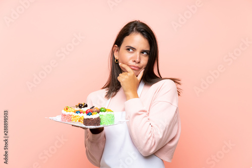 Young girl holding lots of different mini cakes over isolated background thinking an idea