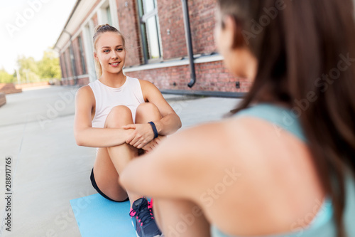 fitness, sport and healthy lifestyle concept - smiling young women or female friends sitting on exercise mats outdoors