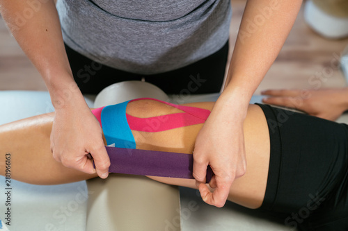 Photo detail of the hands of a physiotherapist woman gluing purple medical tape on another celestial tape and another pink one on the knee of a patient. Concept of muscle health and relaxation.