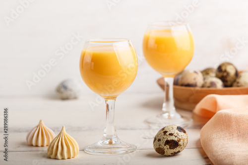 Sweet egg liqueur in glass with quail eggs and meringues on white wooden background. Side view, close up.