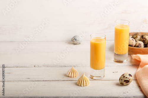 Sweet egg liqueur in glass with quail eggs and meringues on white wooden background. Side view, copy space.