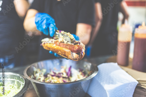 Close up of chef hands adding vegetables on fresh hot dog with grilled sausage.