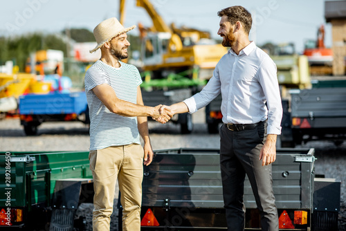 Agronomist shaking hand with salesman, buying a new farm truck trailer on the open ground of the agricultural shop