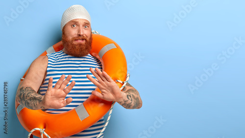 Nervous bearded ginger guy makes refusal gesture, palms outstretched afraids of swimming by himself without help learns swim wears waterproof headgear striped vest carries orange inflated lifebuouy