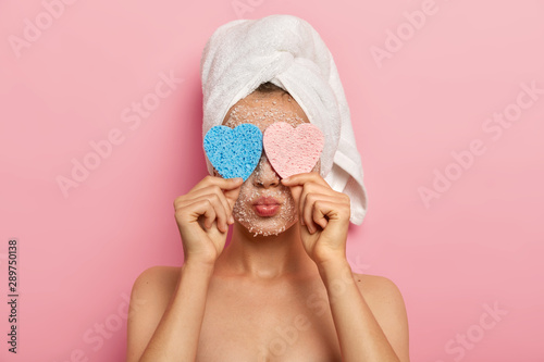 Close up portrait of lovely woman hides face with two cosmetic sponges, has lips folded, wears white soft towel, has naked body, poses over pink background, uses facial scrub for peeling from pores