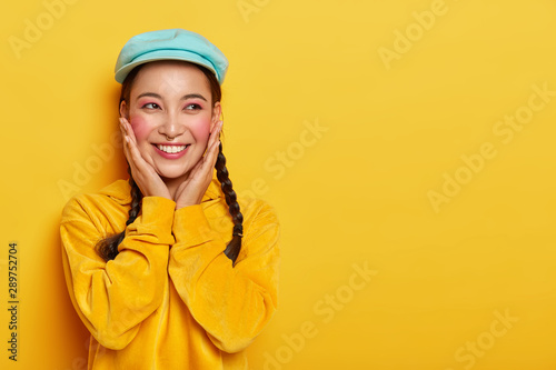 Happy Asian girl touches rouge cheeks, wears makeup and piercing, stylish cap and velvet sweatshirt, has dreamy expression, imagines unforgettable moment with boyfriend, isolated on yellow wall