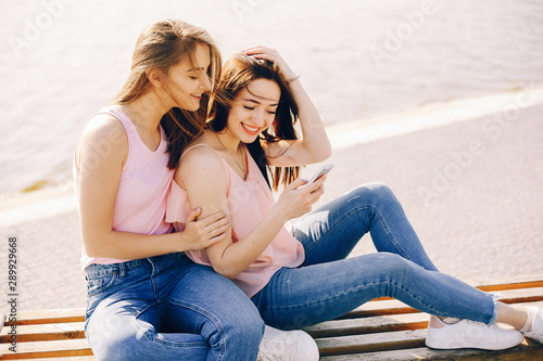 two beautiful and bright friends in pink t-shirts and blue jeans sitting in the sunny summer city and use the phone