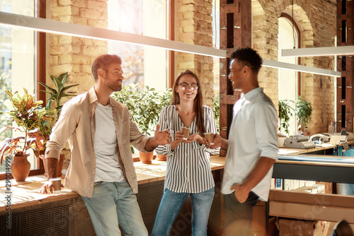 Nice coffee break. Three young and cheerful colleagues in casual wear holding coffee cups and discussing something while standing in the modern office