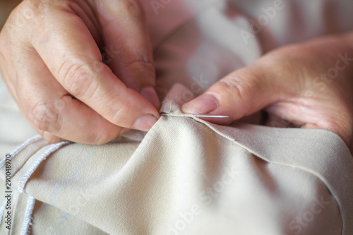 Hand of the seamstress is using a needle to sew brown pants by hand close-up.