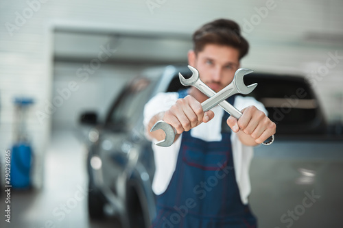 young handsome mechanic in uniform holding two spanners crossed stands in car service center
