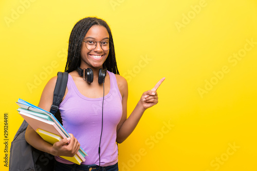 African American teenager student girl with long braided hair over isolated yellow wall pointing finger to the side
