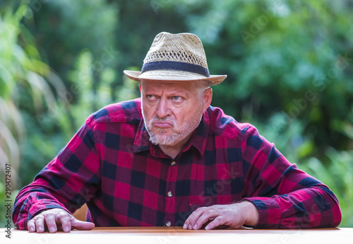 An elderly man in a hat is sitting with bad mood at a table in the garden. Concept: evil and unsympathetic people
