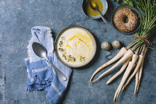 Parsnipcream soup in ceramic bowl with butter sauce, bagel bread, bundle of fresh parsnip and herbs over blue texture background. Flat lay, space