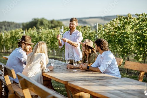 Group of a young people drinking wine and talking together while sitting at the dining table outdoors on the vineyard on a sunny evening