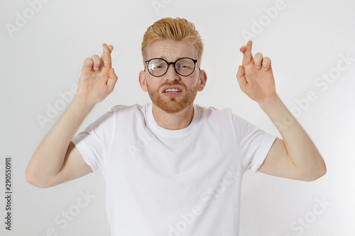 Fingers crossed, make a wish concept. Young redheaded guy with red beard cross finger for good luck. Hand sign, gesture for dreams come true. Man in blank template white t shirt. Mockup, copy space