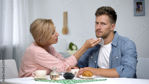 Mother wiping irritated adult sons mouth during having tea at home, care