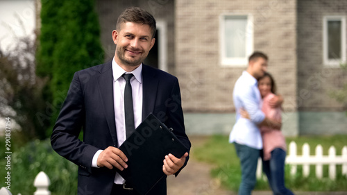 Smiling broker standing with documents, happy family hugging near their new home