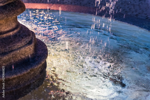 Closeup of splashing water fountain in downtown village park during evening dark night with illuminated light lamps