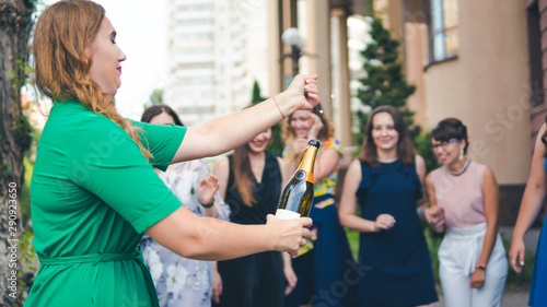 Young woman opening a bottle of champagne with female friends standing in background laughing and having fun. Young people celebrating with champagne at party outdoors.