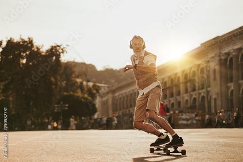 Portrait of handsome man in white shirt on sunset evening in summer. Stylish guy ride on skateboard on city street. Urban male lifestyle on buildings background
