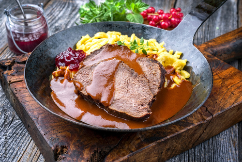 Traditioneller geschmorter marinierter deutscher Sauerbraten vom Rind mit Spätzle und Preiselbeer Marmelade in in würziger brauner Sauce als closeup in einer rustikalen gußeisernen Bratpfanne