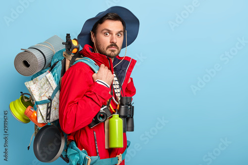 Photo of male vacationist has active rest, carries rucksack with map, rolled up rag, wears casual tourist clothes, uses binoculars, flask with hot drink surprised by something. Backpacking concept